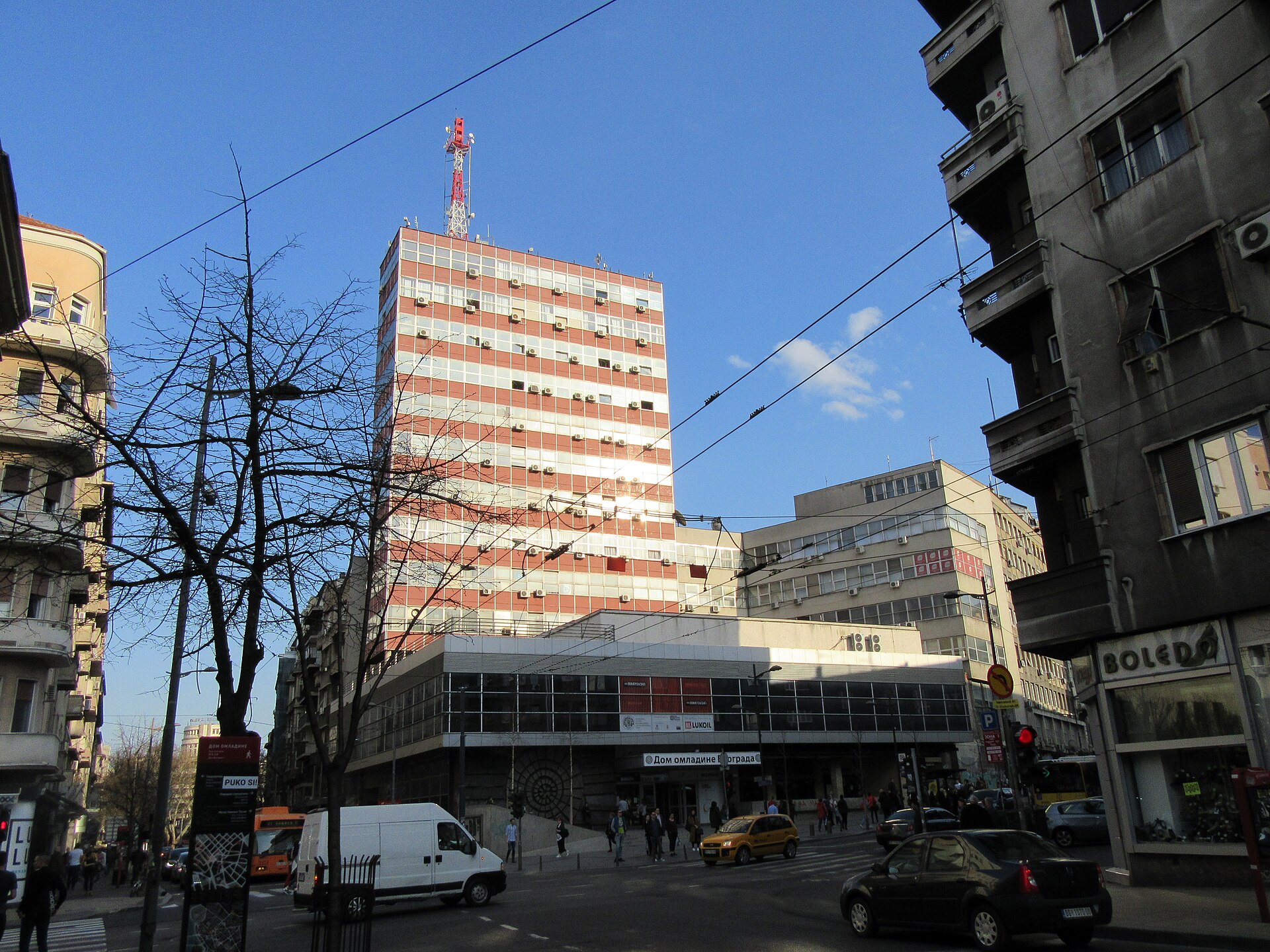 Belgrade street intersection with mid-rise buildings, tram wires, and Dom omladine beneath a striped tower