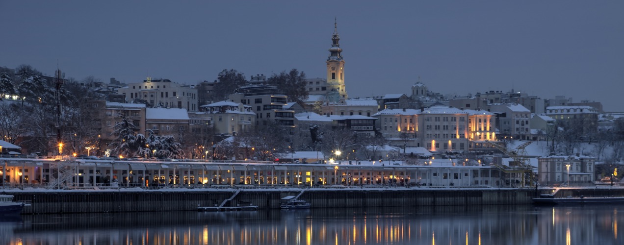 Snowy Belgrade riverfront with lit buildings and reflections on the water at dusk