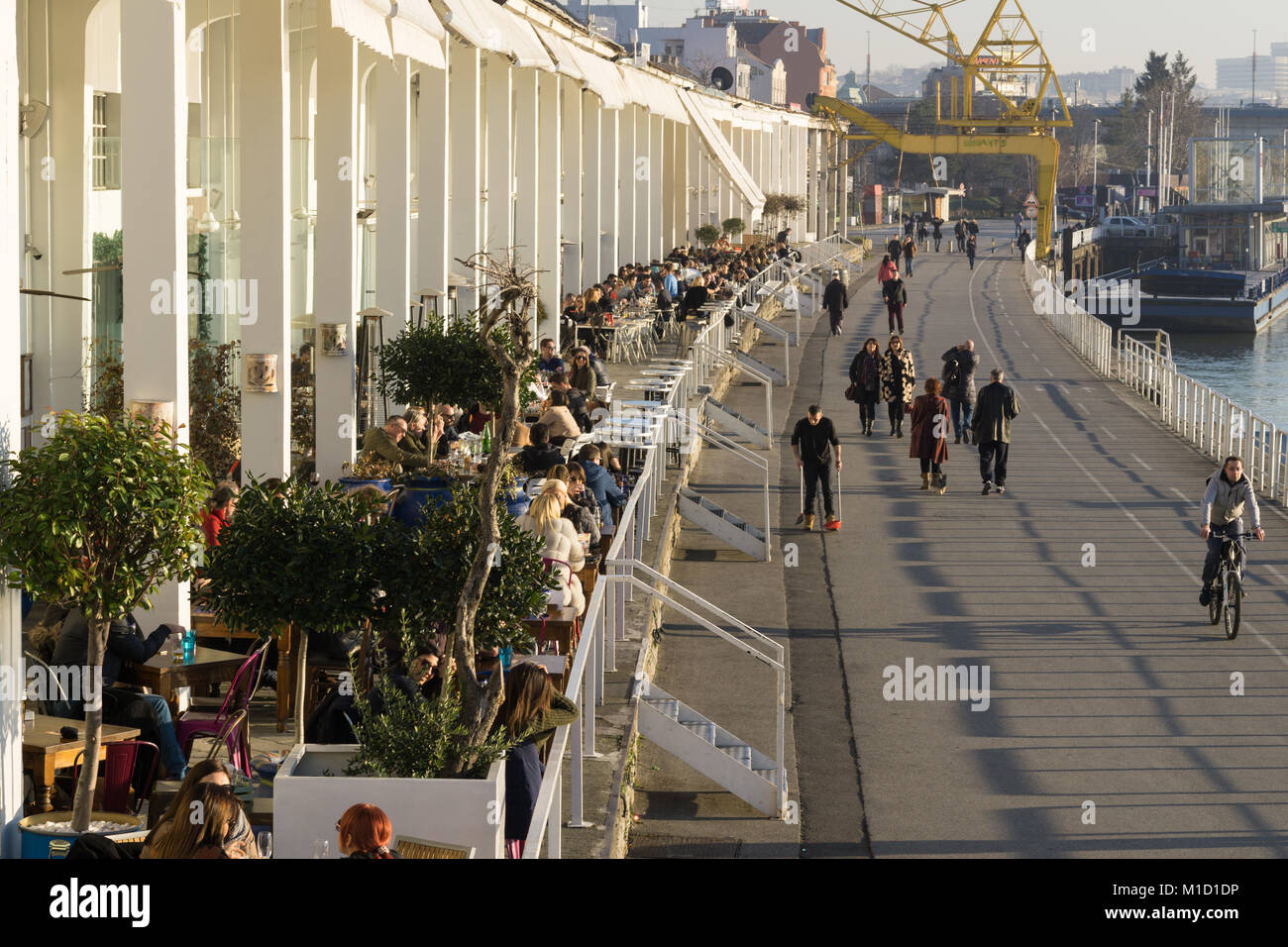 Outdoor cafes and a riverside promenade at Beton Hala, Belgrade, with pedestrians, cyclists, and cranes