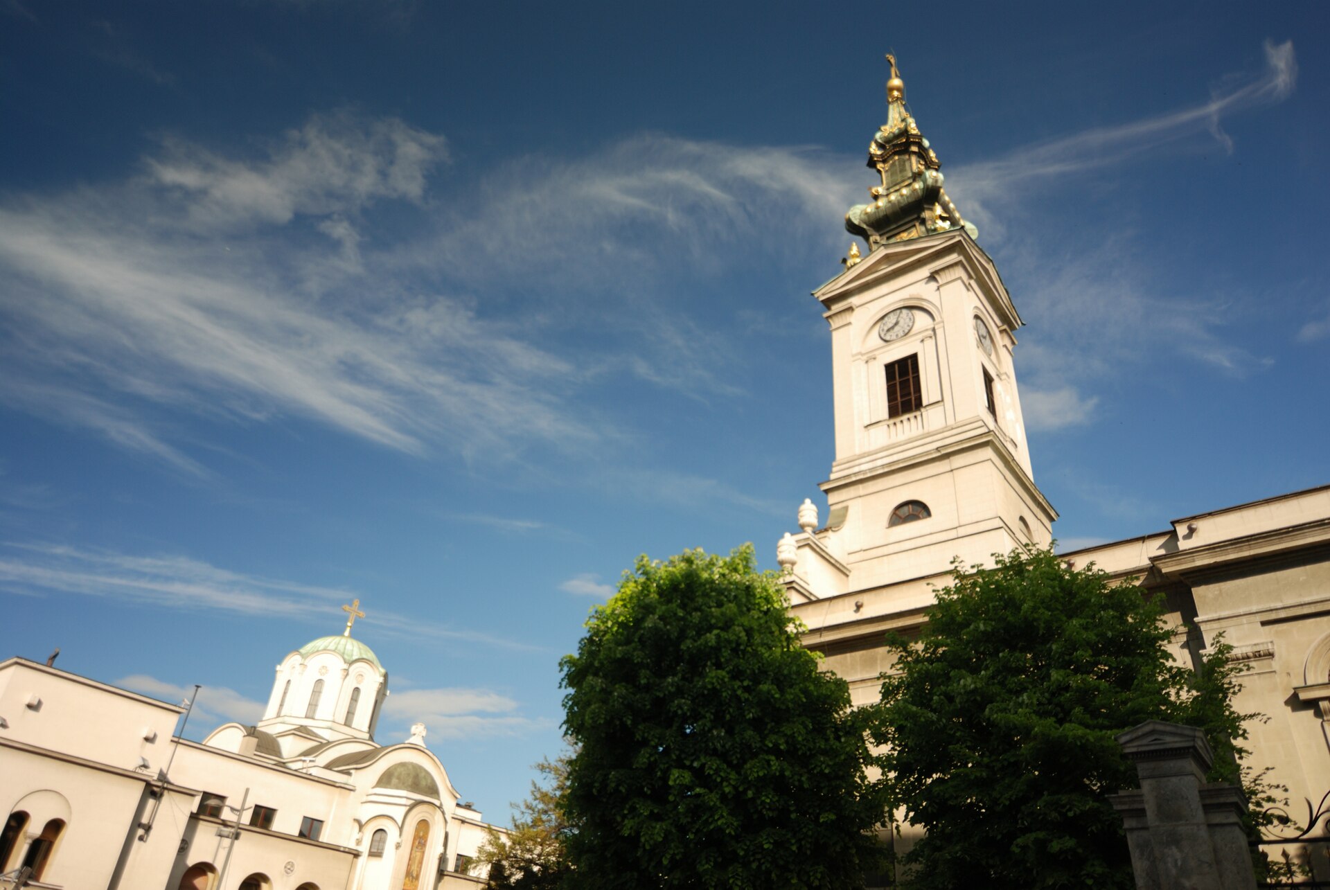 Cathedral of St. Michael in Belgrade with a clock tower, green dome, and trees under a blue sky