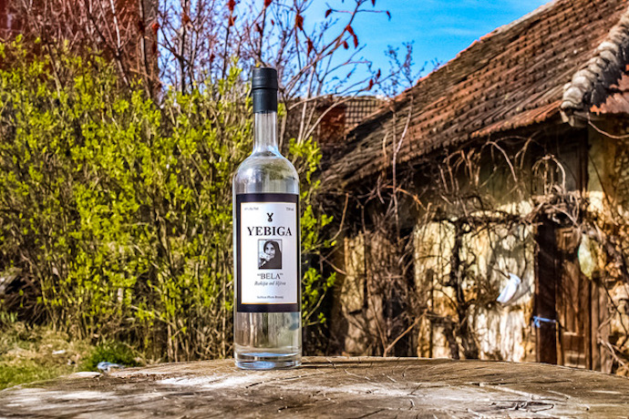 Yebiga rakija bottle on a wooden table with a rustic house behind it in Rakija