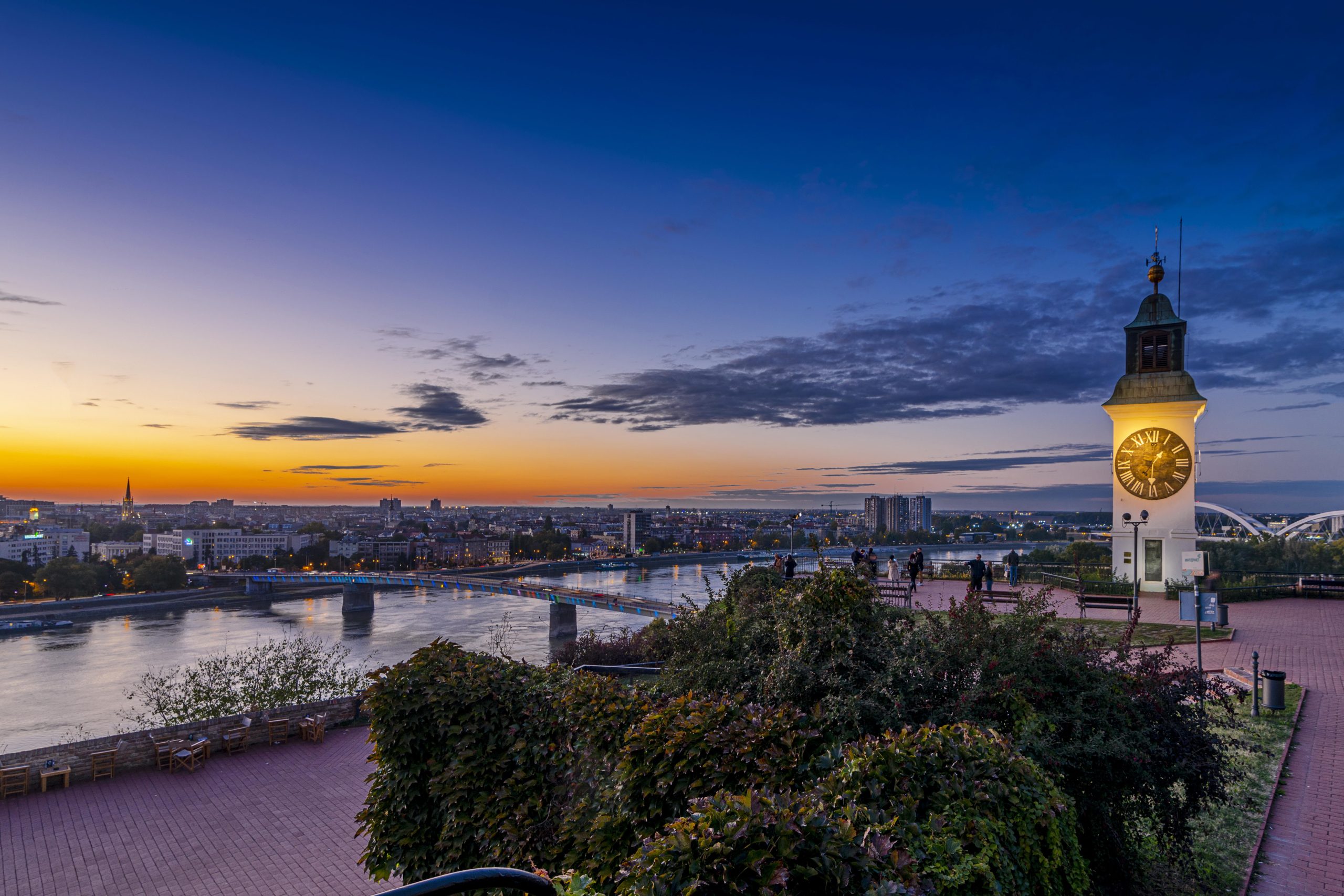 Clock tower above the Danube at sunset, with Novi Sad skyline and a bridge nearby