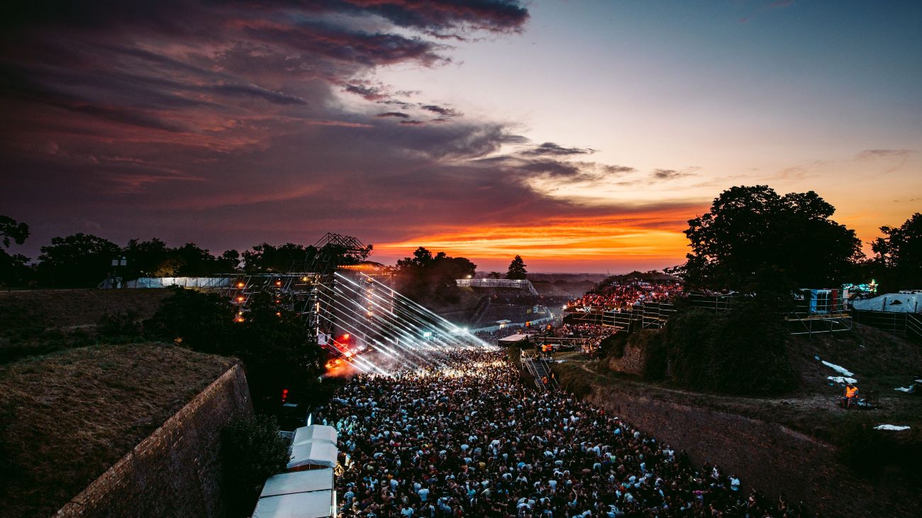 Crowd gathered before a brightly lit concert stage at sunset inside the fortress in Novi Sad