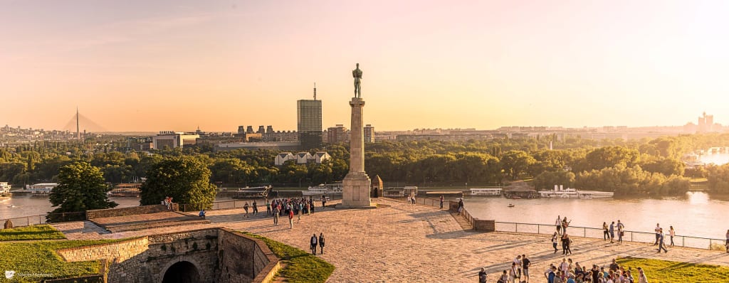Stone terrace with the Victor monument above the river and visitors at Belgrade Fortress at sunset