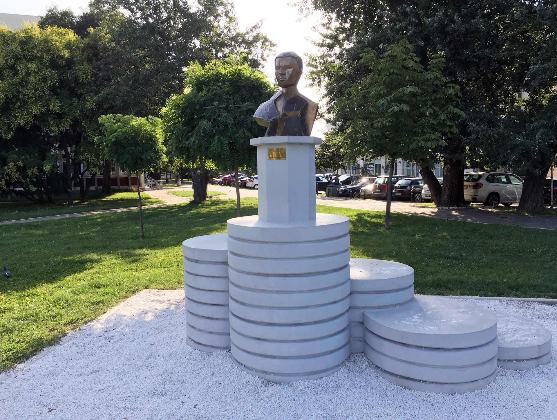 Geometric bust sculpture on a stepped pedestal in Freedom Square, Novi Sad, with trees and grass nearby