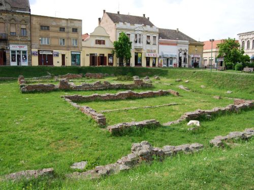 Low brick ruins in grass beside buildings at the Imperial Palace of Sirmium