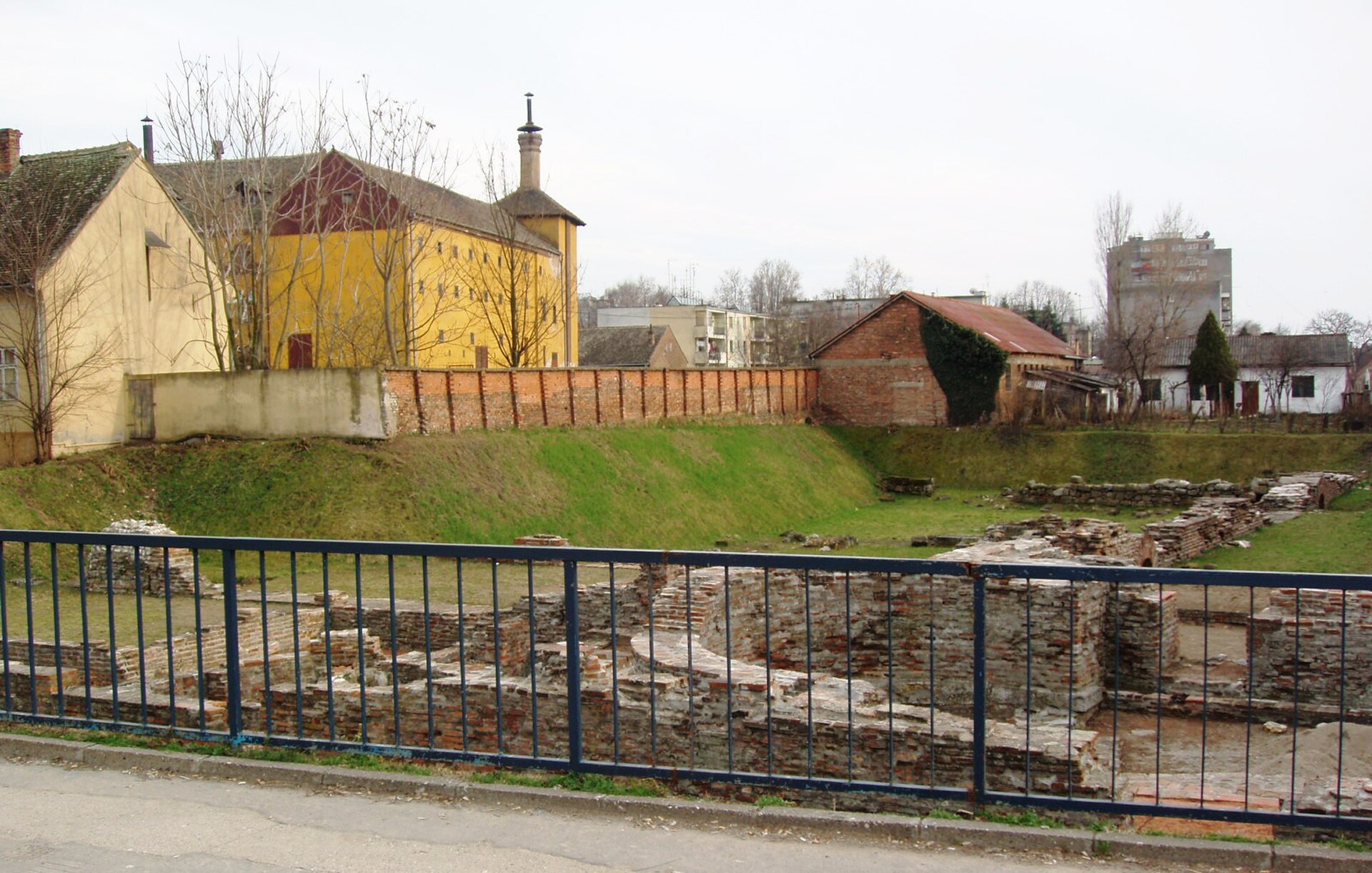 Brick ruins and grassy foundations behind a blue fence at the Imperial Palace of Sirmium
