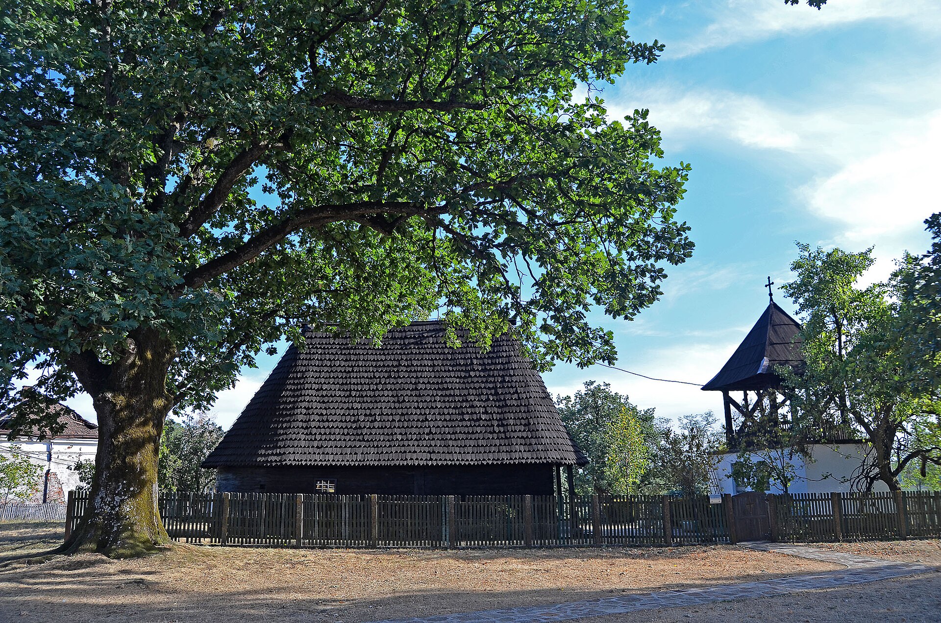 Fenced wooden church with bell tower under large trees in a Serbian destination setting