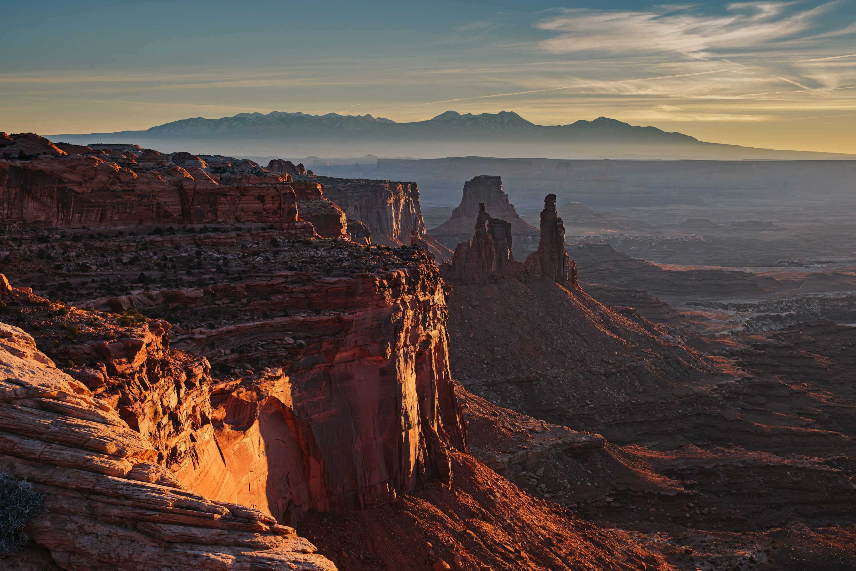 Sunlit red sandstone cliffs and spires overlooking a hazy desert valley and distant mountains