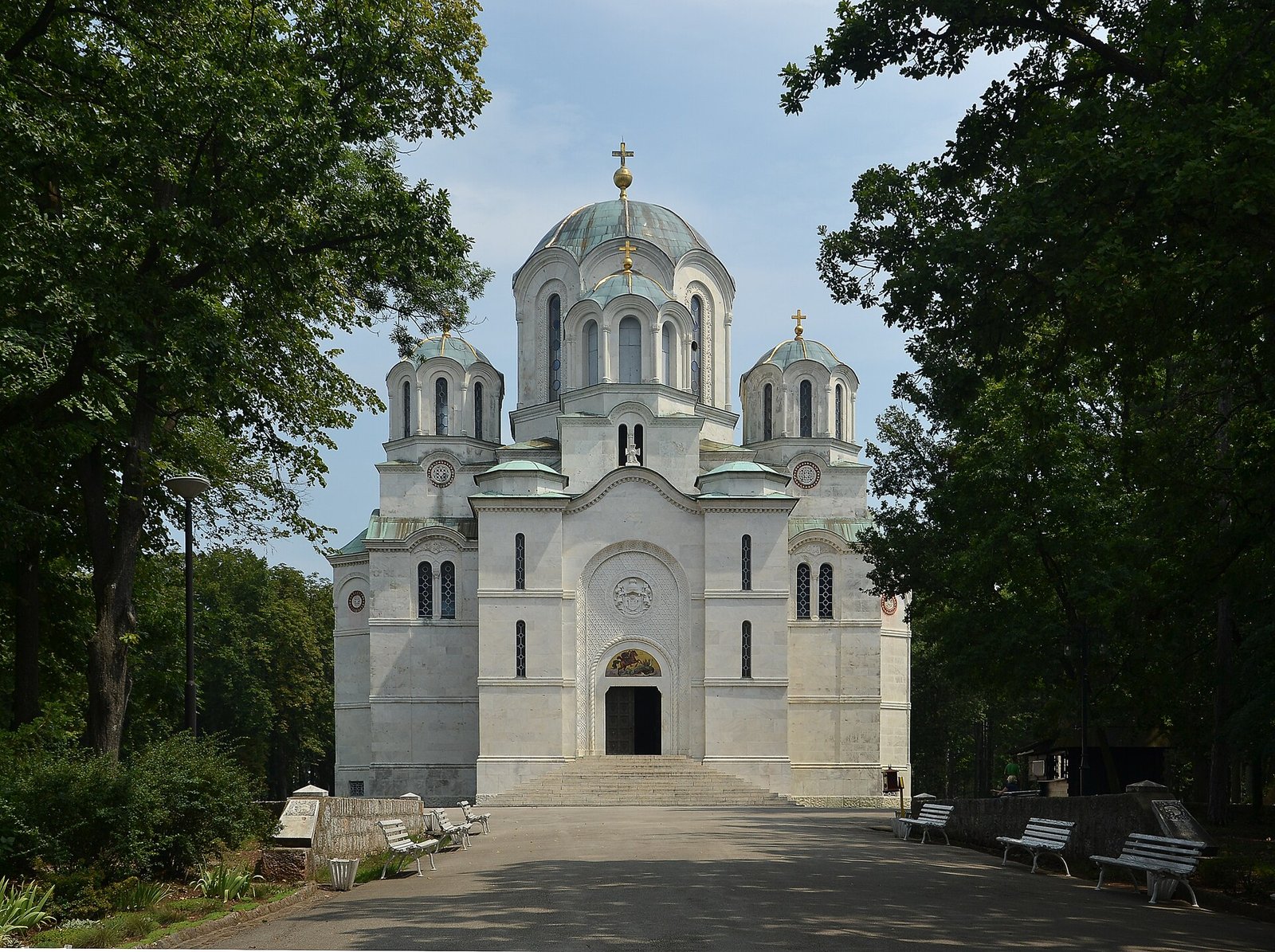 White Orthodox church with domes and crosses, framed by trees and benches in Kragujevac
