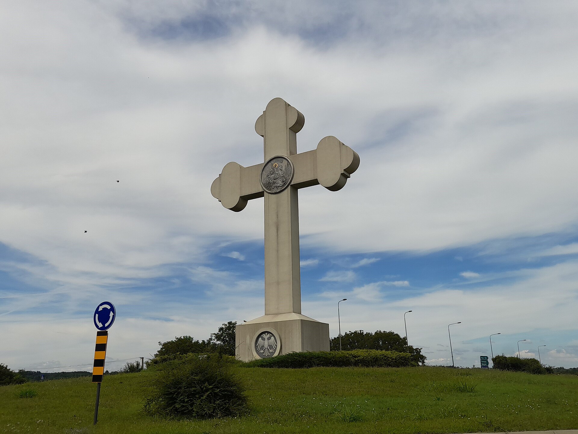 Tall stone cross monument on a grassy roundabout in Kragujevac under a cloudy sky