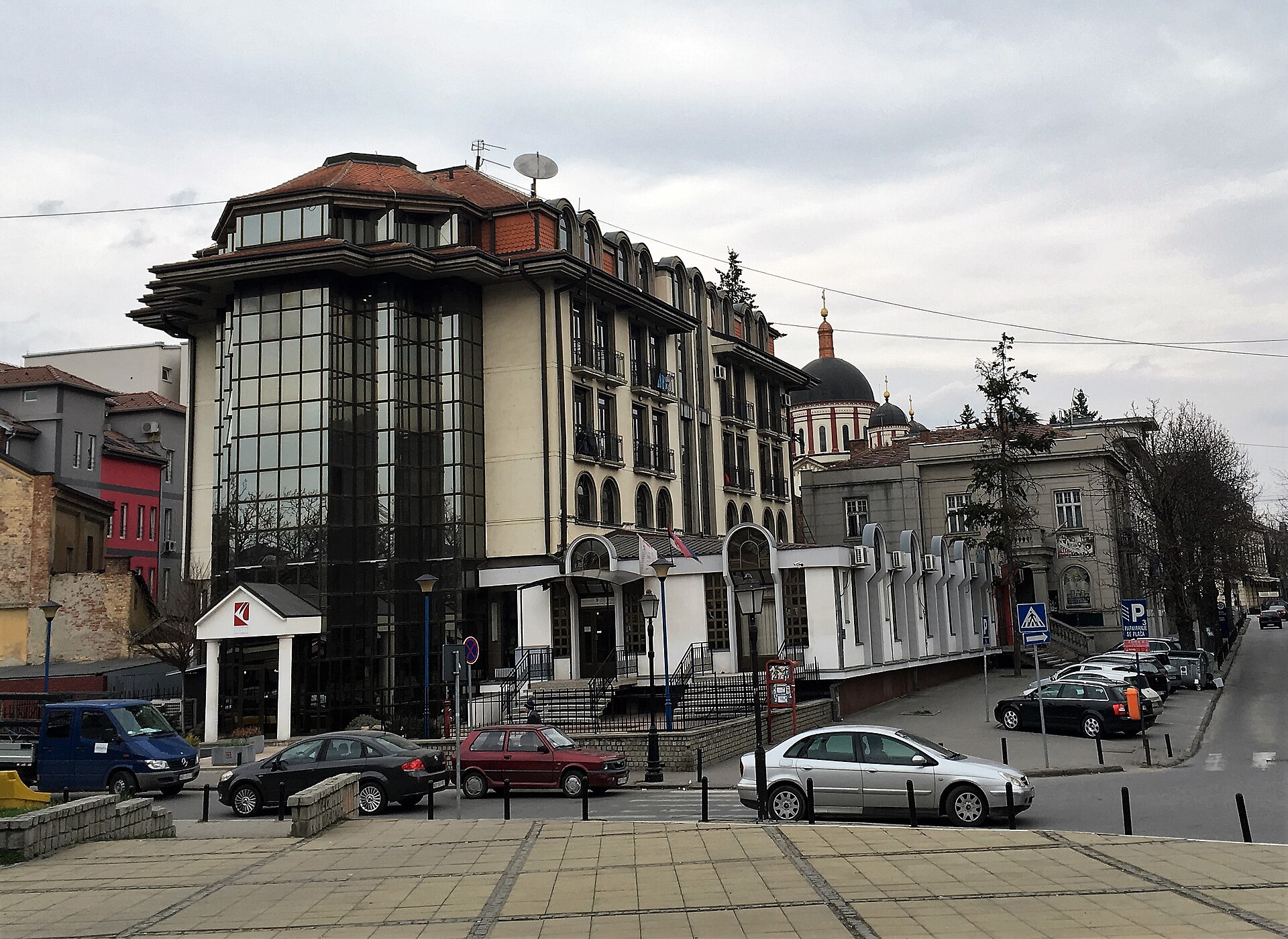Modern corner building with glass facade, parked cars, and church domes behind in Kragujevac