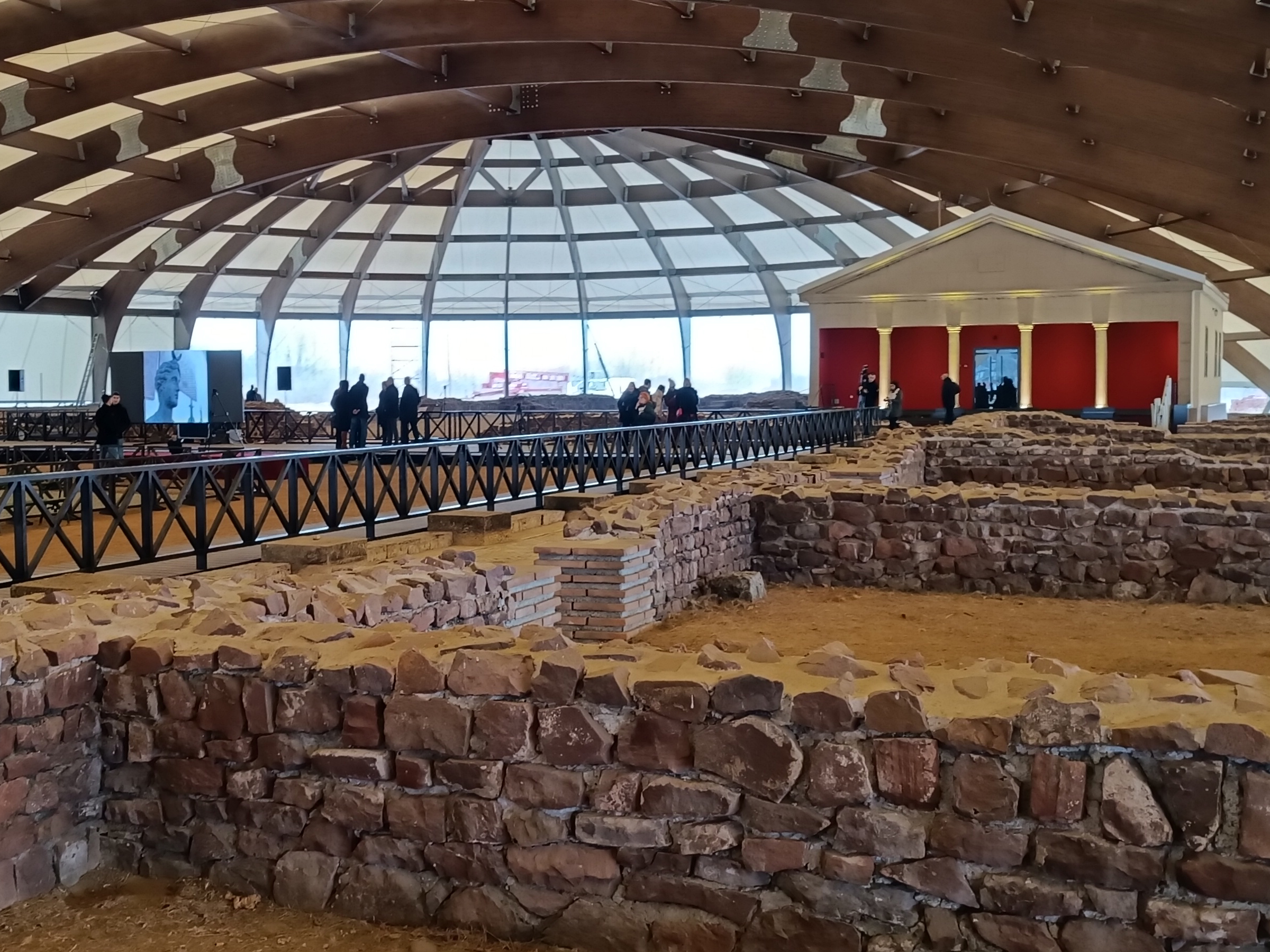 Stone ruins beneath a large dome with visitors and a red exhibition building at Mediana in Niš