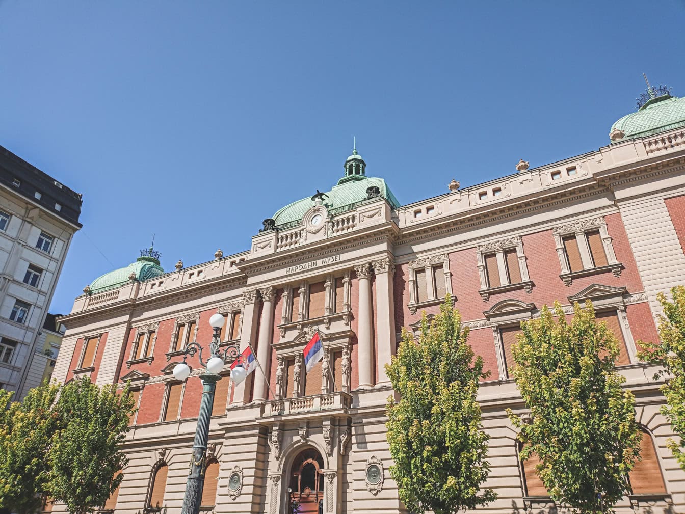 Neoclassical facade of the National Museum in Belgrade with green domes, columns, and Serbian flags