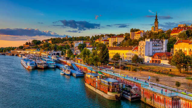 Riverboats moored beside a waterfront promenade with hillside buildings and a church tower in Novi Beograd