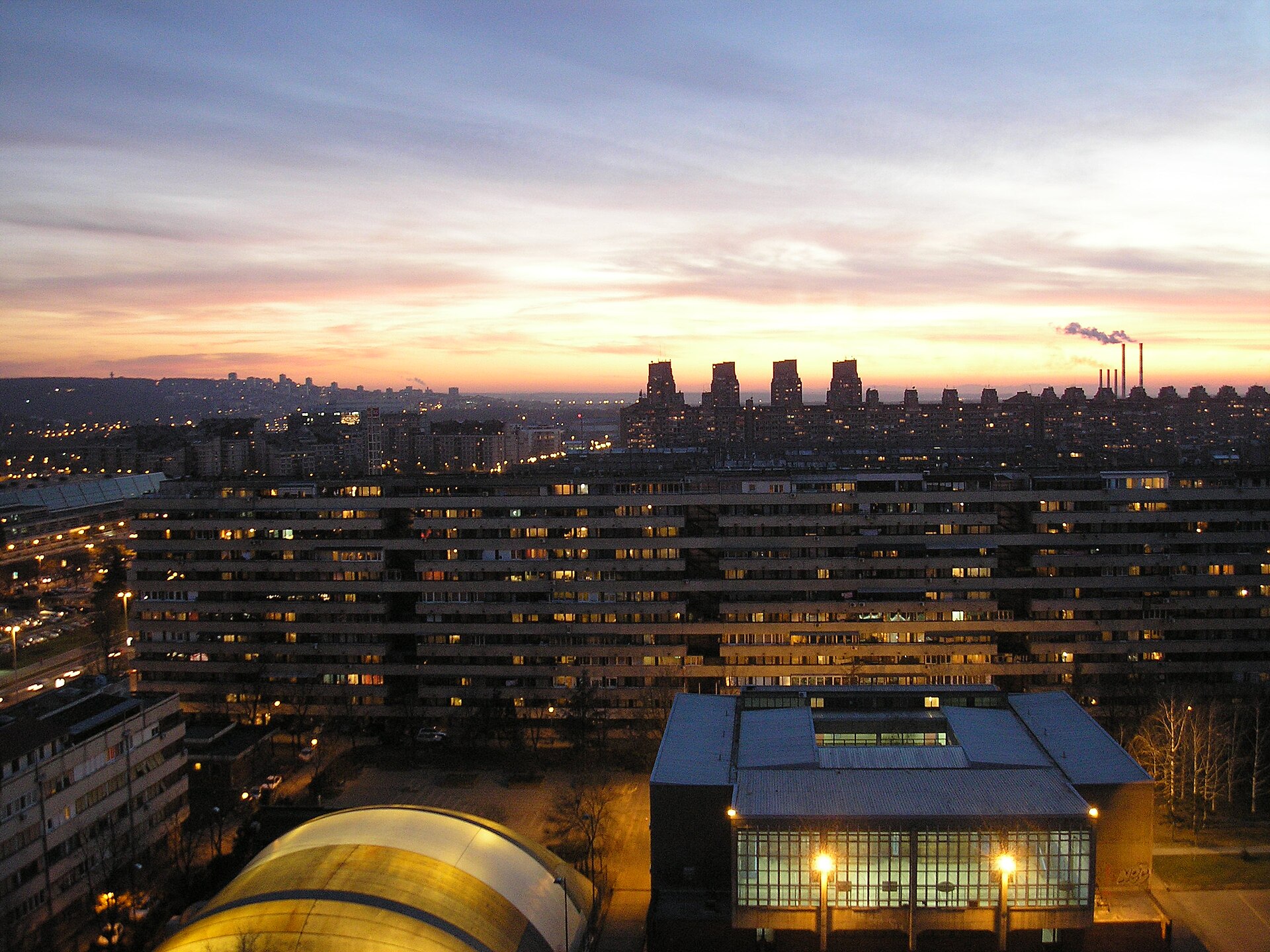 Dusk skyline over Novi Beograd with apartment blocks, lit windows, and smokestacks under a colorful sky