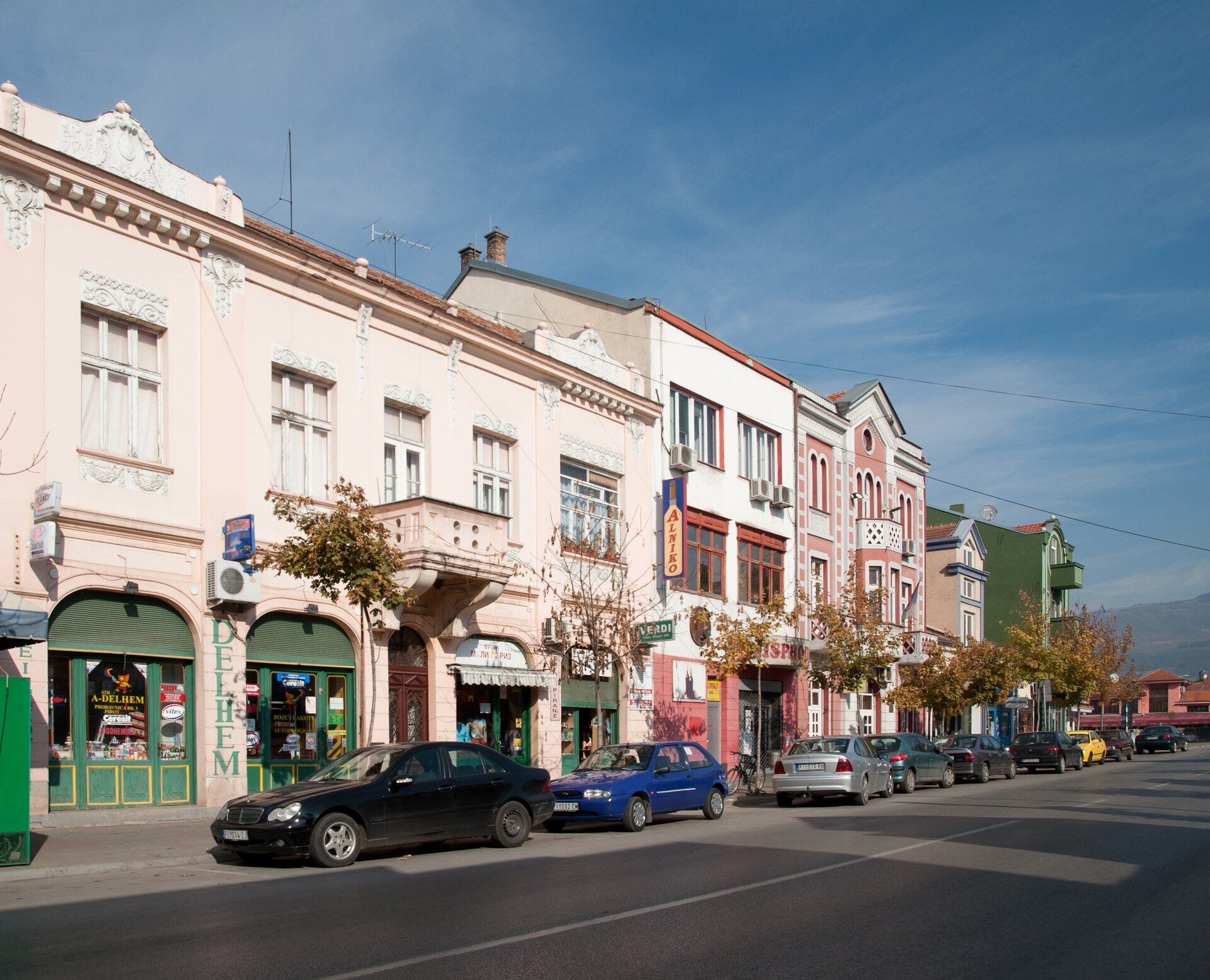 Pastel historic buildings with shopfronts and parked cars along a street in Pirot