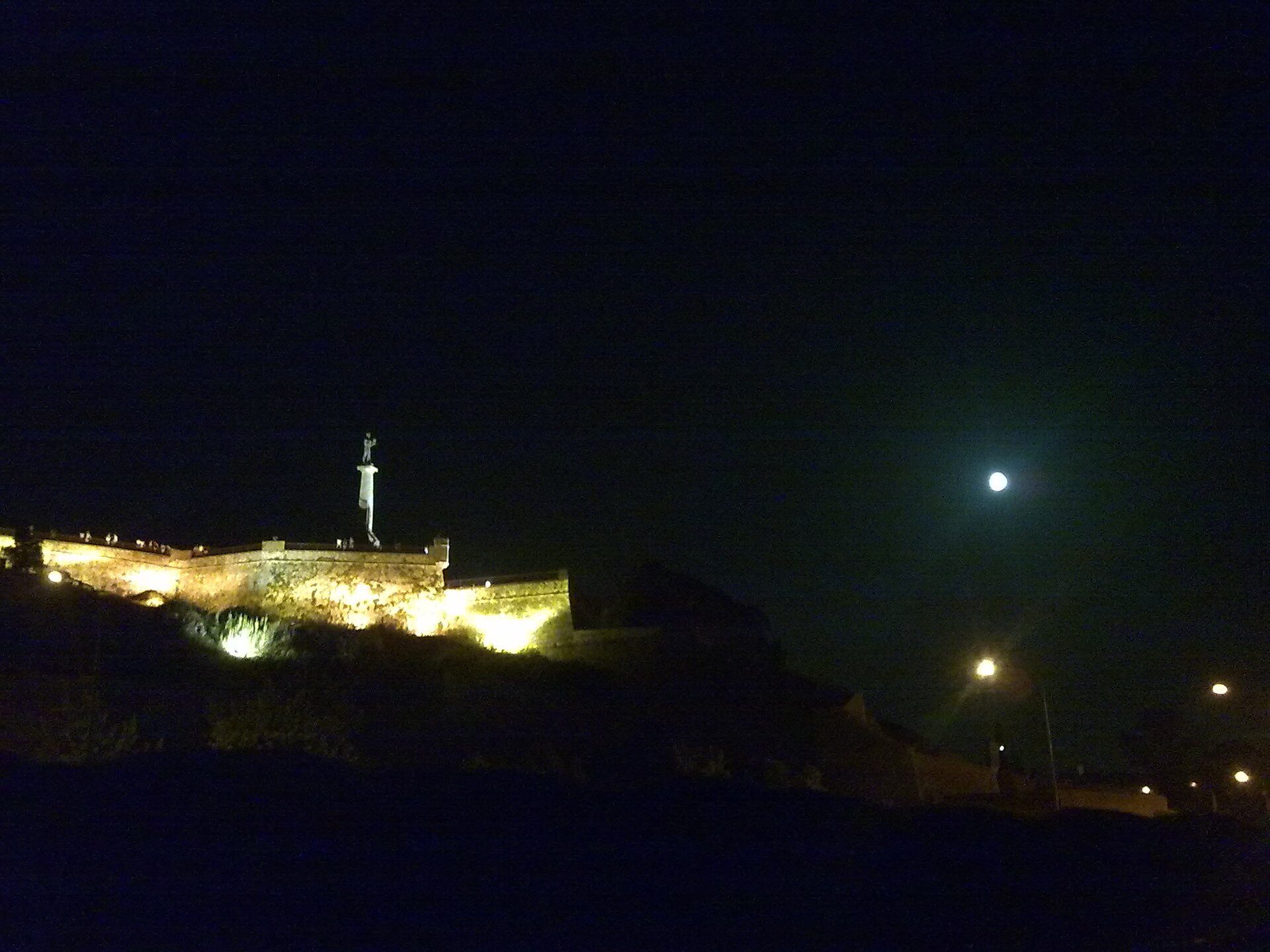 Pobednik Monument above lit fortress walls at night in Belgrade, with the moon in the sky