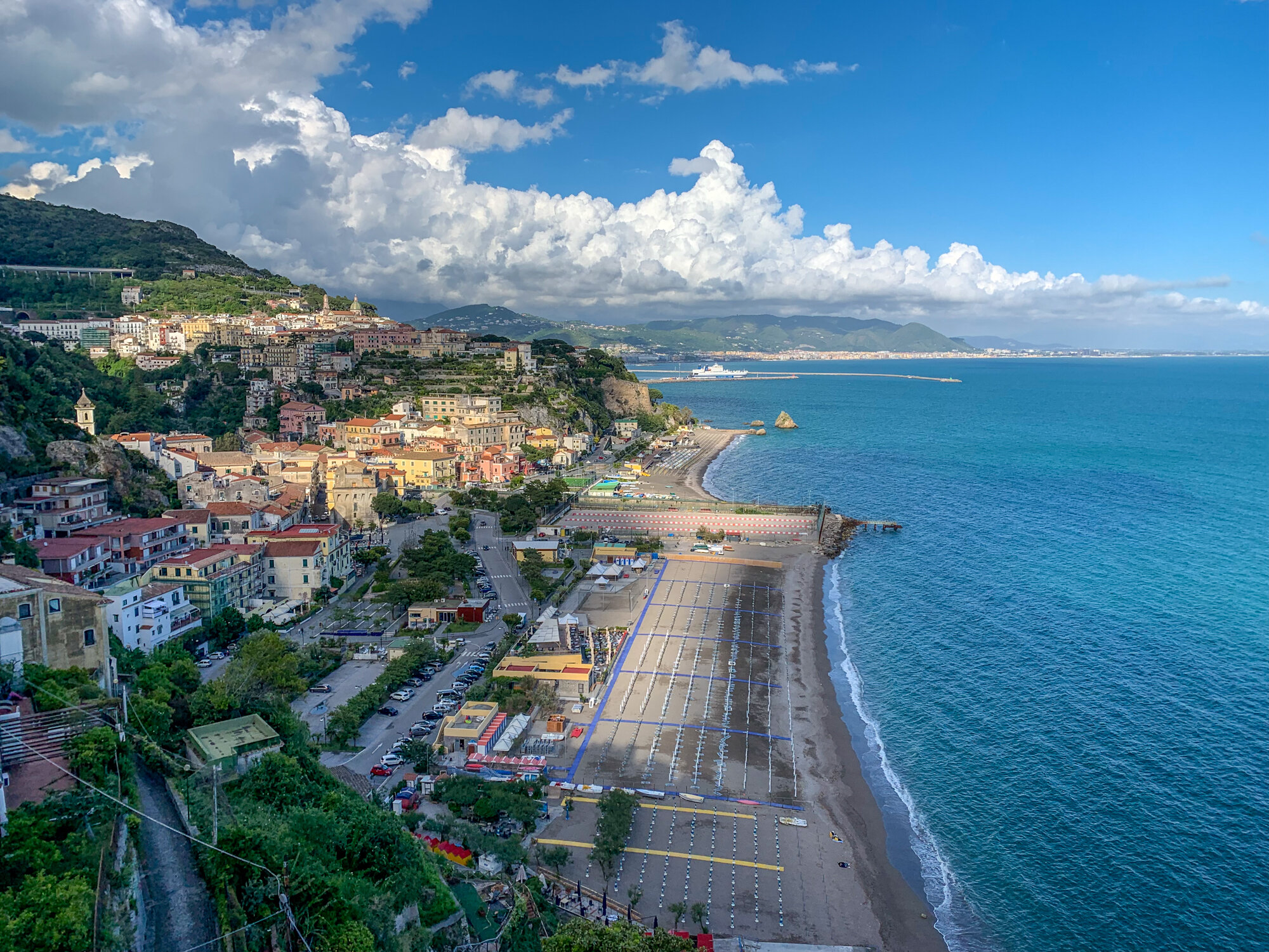 Hillside town above a long beach and blue coastline with scattered clouds overhead