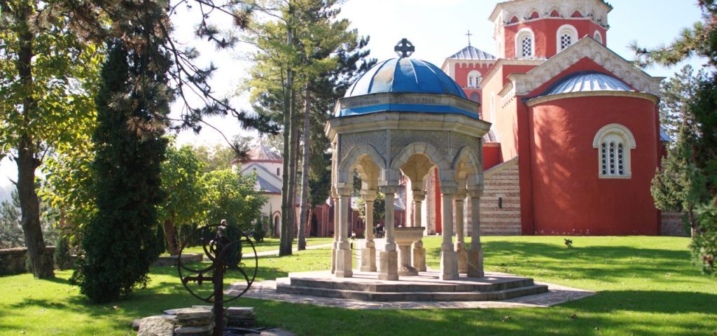Stone pavilion in a grassy Serbian monastery courtyard with red domed church buildings behind it