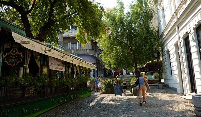 Cobblestone street with outdoor café seating, trees, and pedestrians in Skadarlija Bohemian Quarter, Belgrade