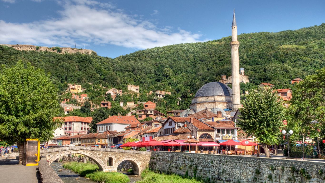 Stone bridge over a narrow river beside a mosque, red-roofed buildings, and green hills in Sombor