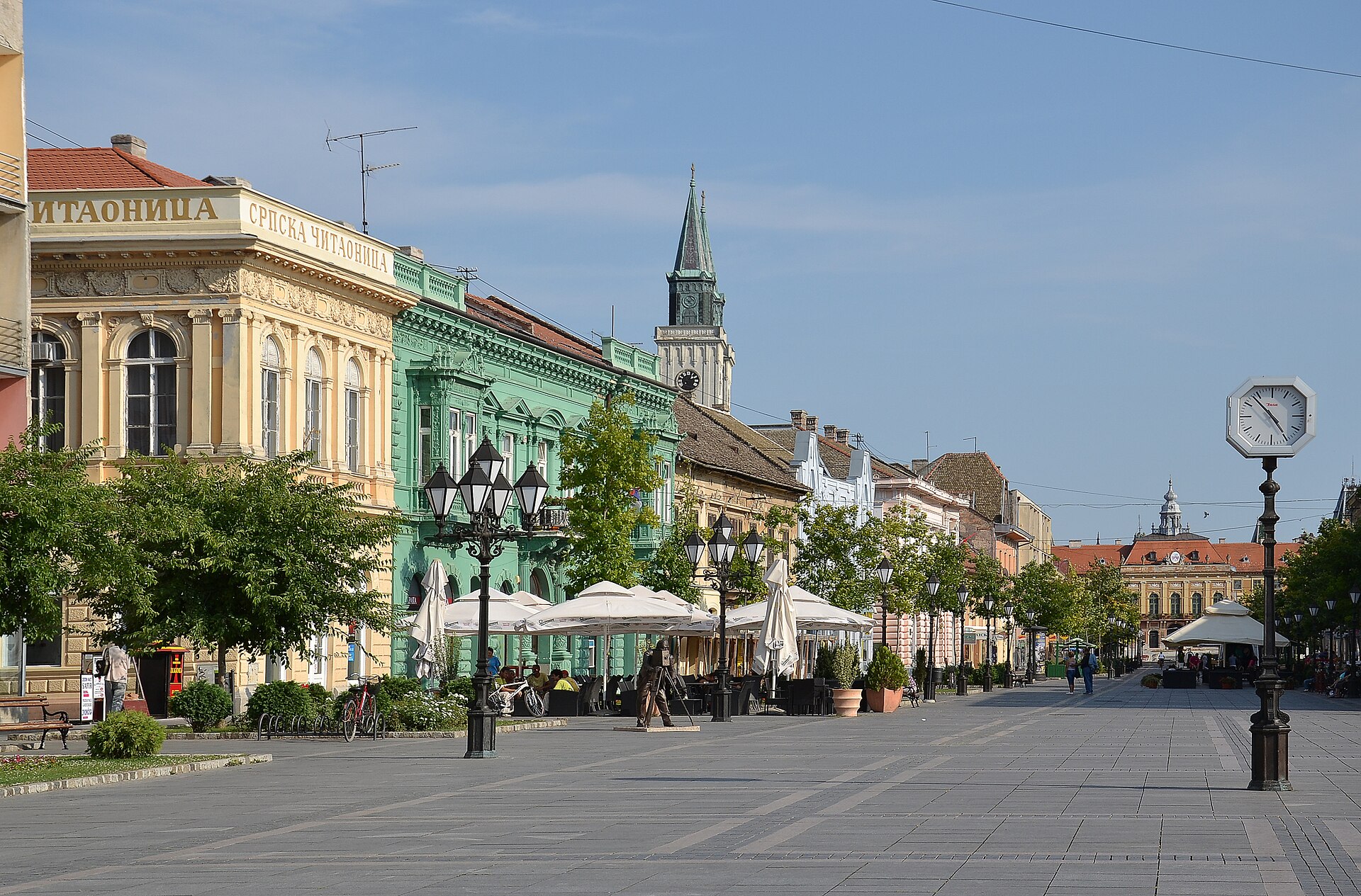 Pedestrian street with colorful historic buildings, café terraces, and a church tower in Sombor