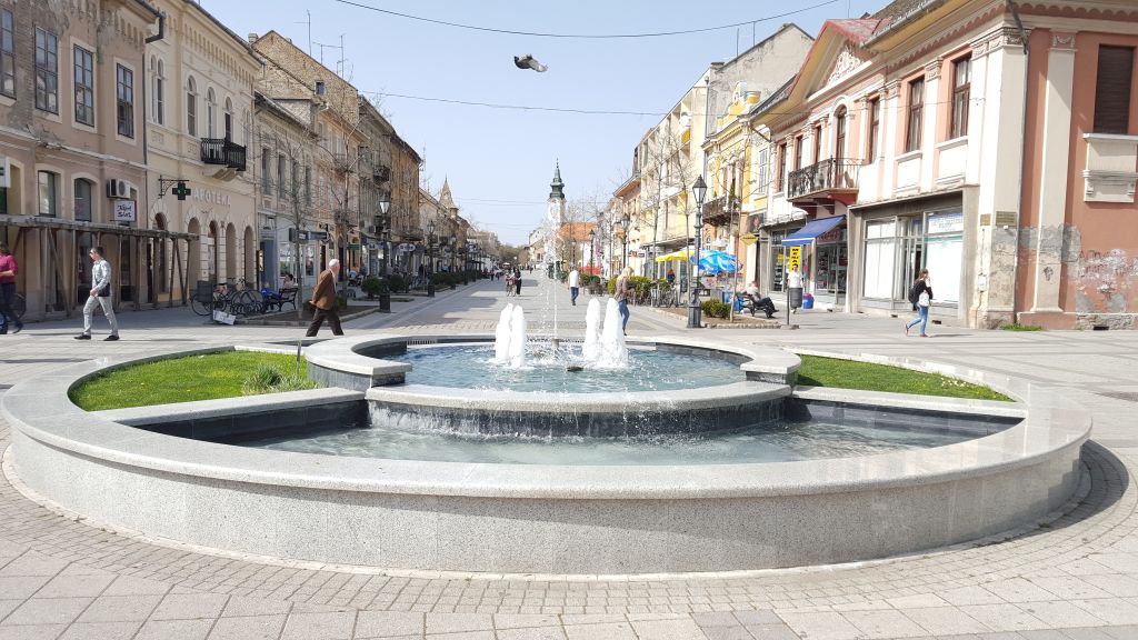 Circular fountain in a pedestrian street lined with pastel buildings in Sombor