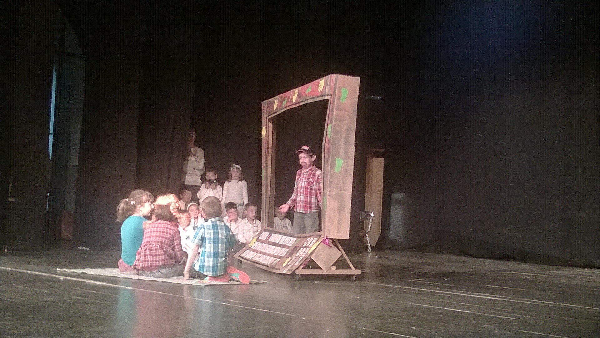 Children sit on a stage watching a man standing inside a decorated cardboard frame