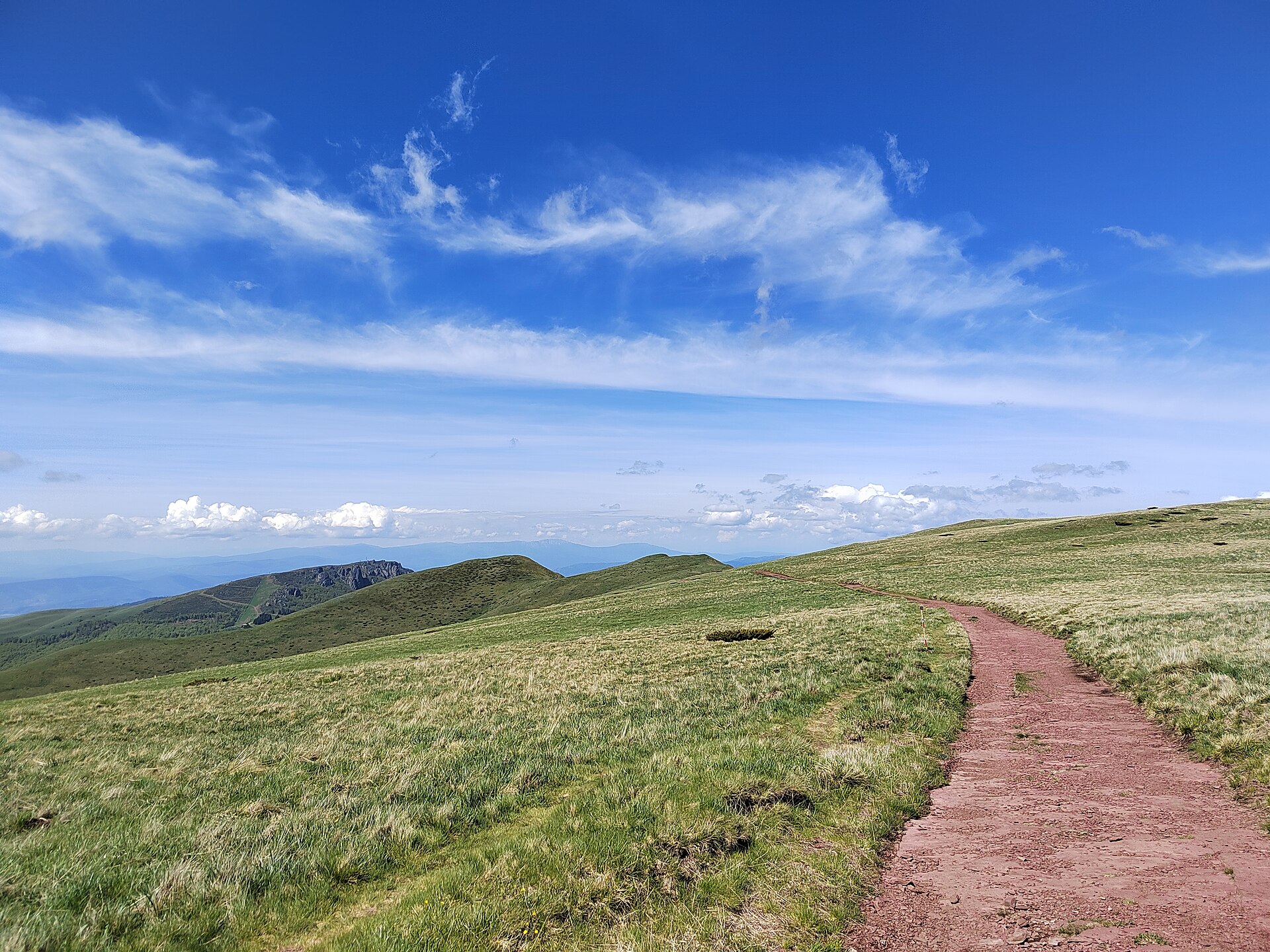 Red dirt trail winding across grassy alpine hills beneath a wide blue sky in Stara Planina