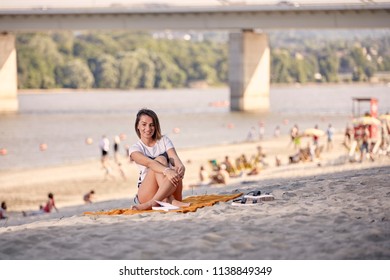 Woman sitting on a sandy river beach with swimmers and a bridge across the water in Djerdap National Park