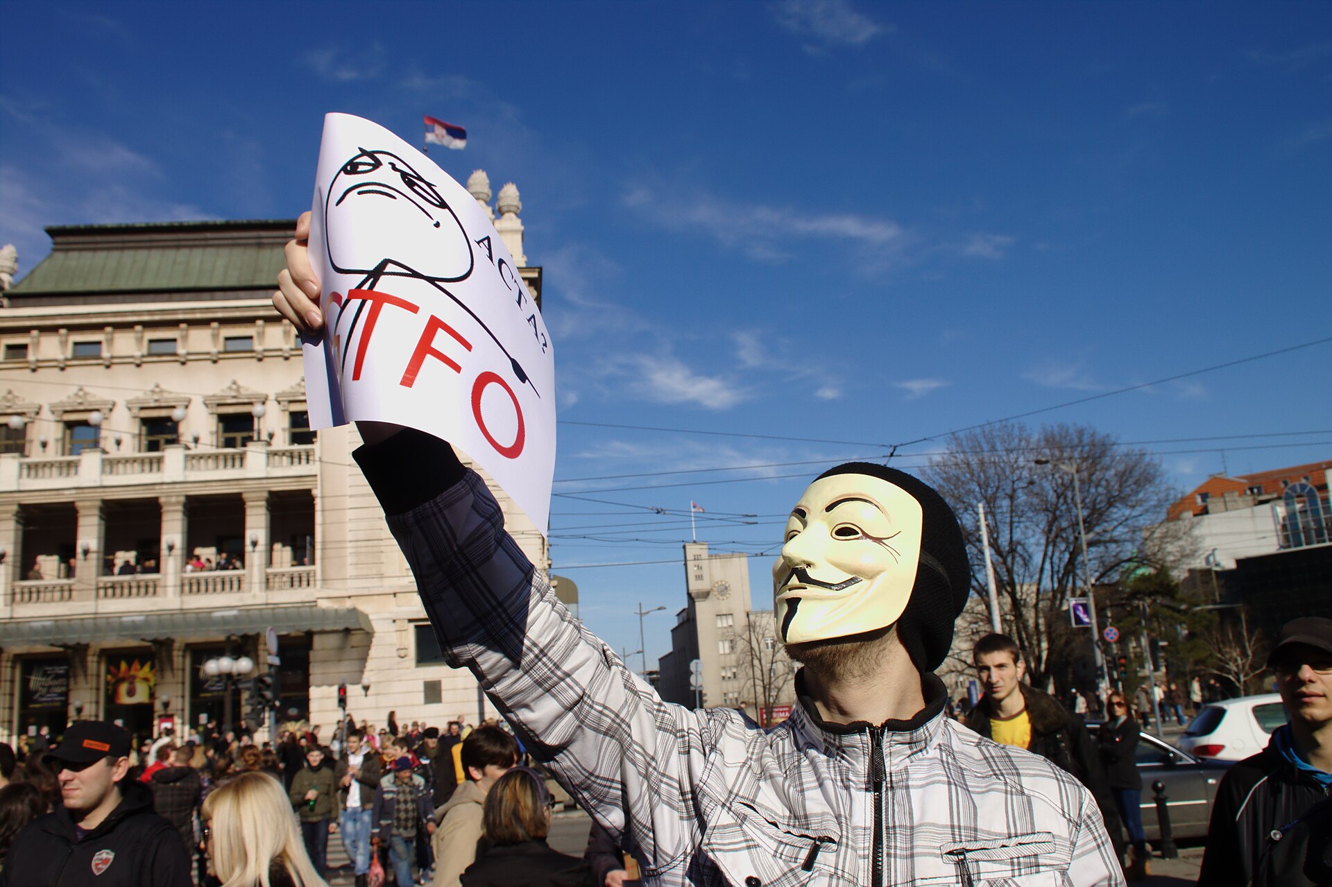 Masked protester holding a sign in a crowd at Trg Republike, Belgrade