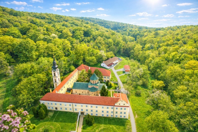 A monastery complex with red roofs and a bell tower surrounded by dense green forest in Vinča
