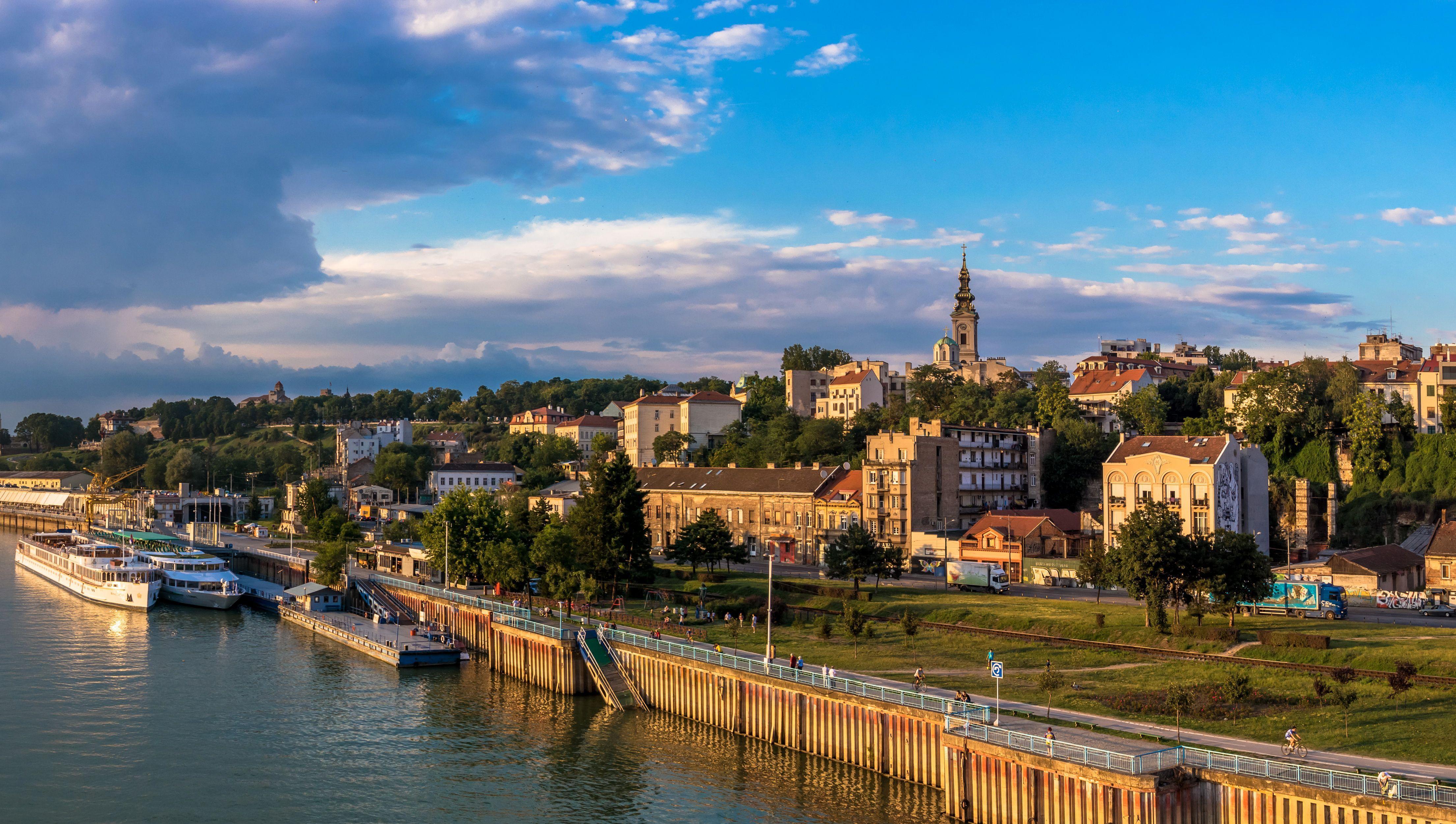 Belgrade riverfront with docked boats, embankment walkway, and hillside buildings under a cloudy sky
