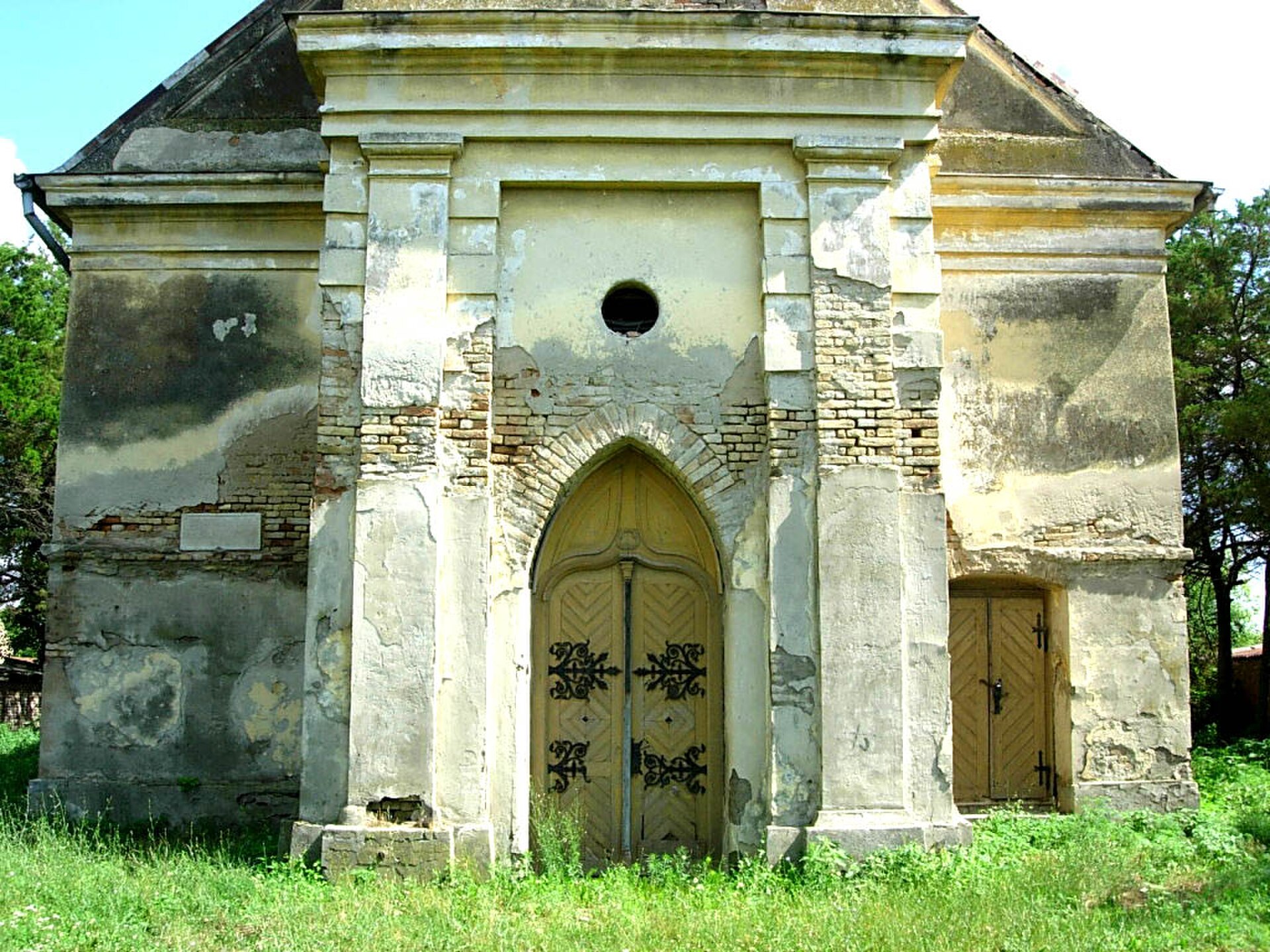 Weathered chapel with arched wooden doors and peeling plaster in Vojvodina, surrounded by grass