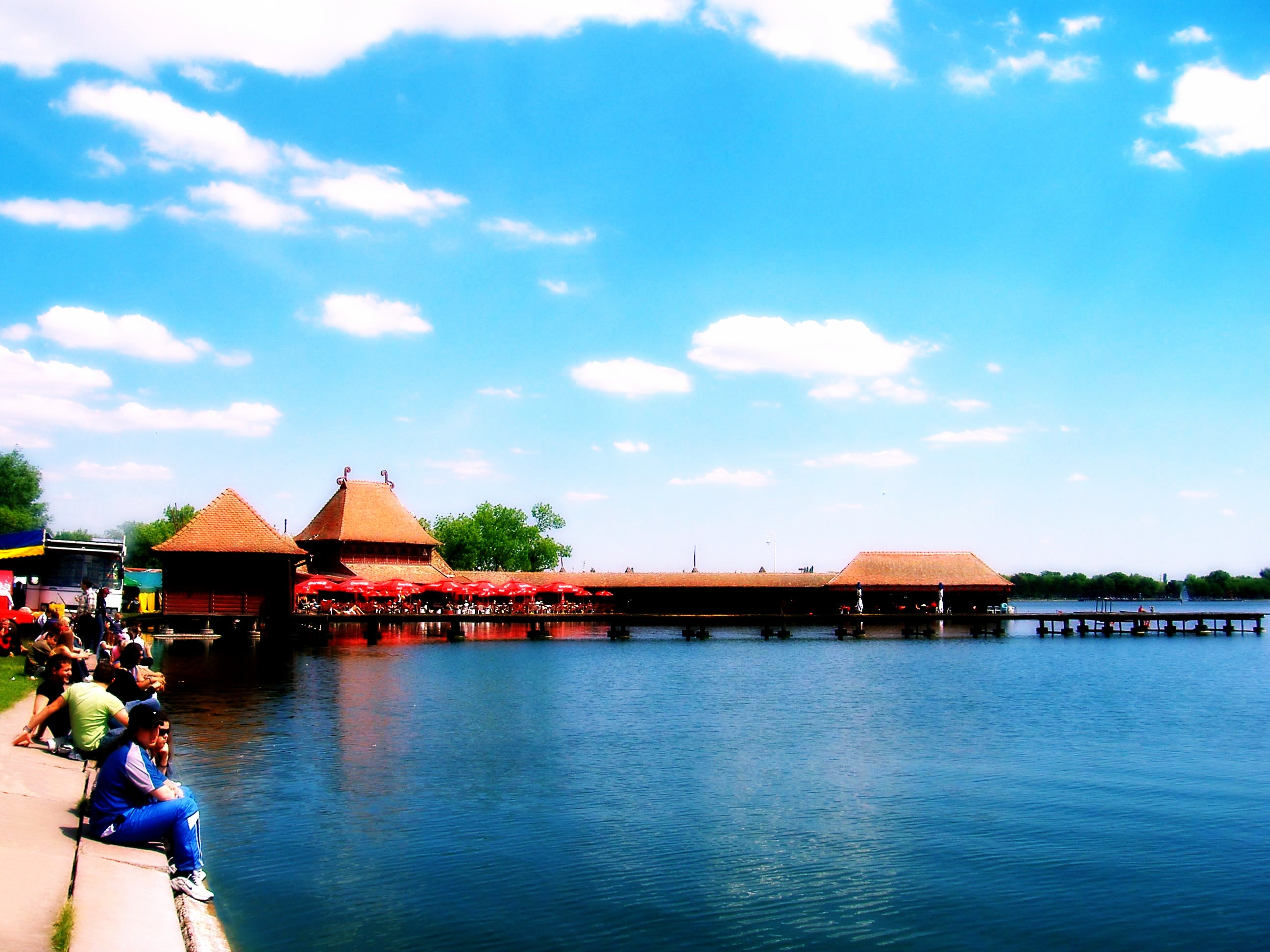People sitting by a lakeside boardwalk with thatched pavilions and red umbrellas in Vojvodina