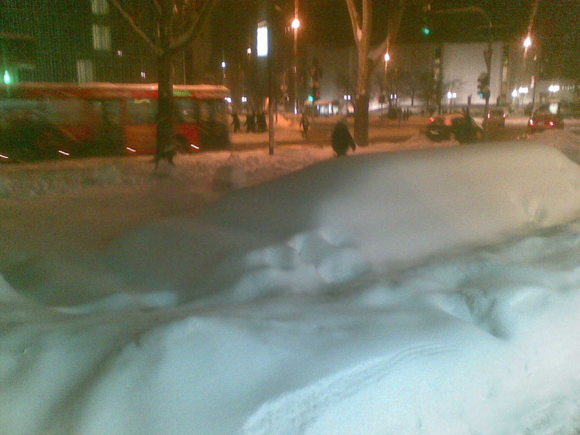 Night street in Novi Sad with a tram, pedestrians, and deep snowbanks under streetlights