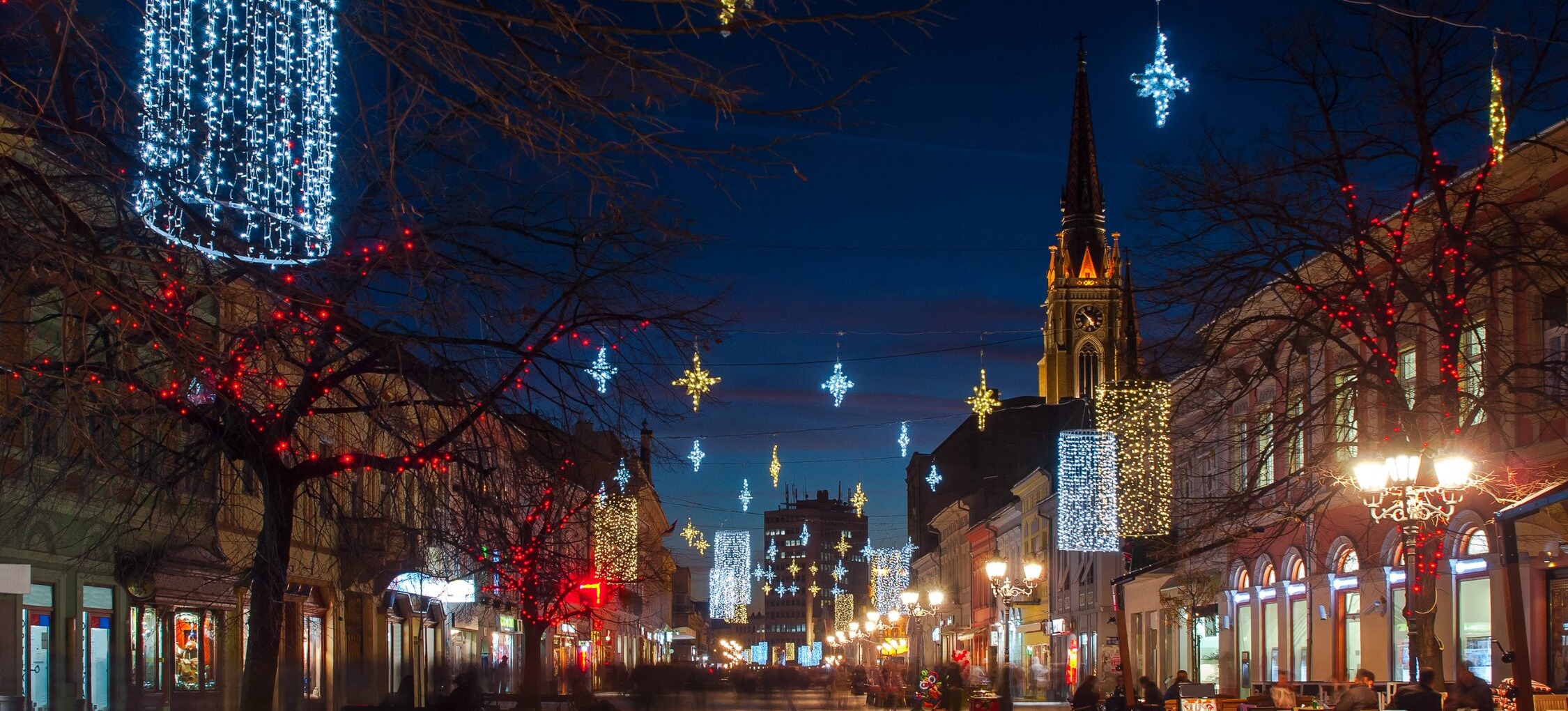 Festive lights hang over a pedestrian street in Novi Sad, leading toward a church spire at dusk