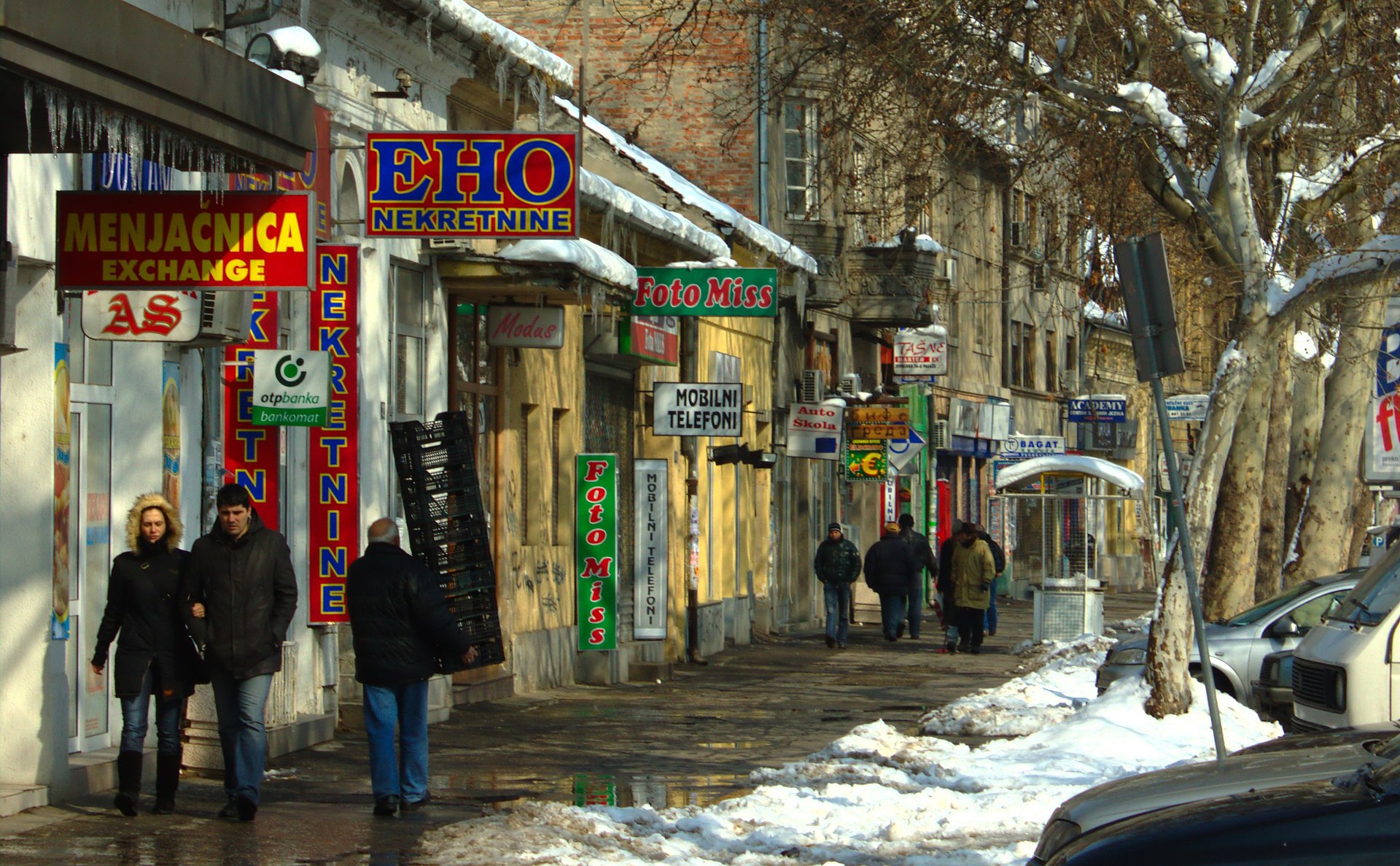 Snowy sidewalk with storefront signs, pedestrians, and parked cars in Novi Sad