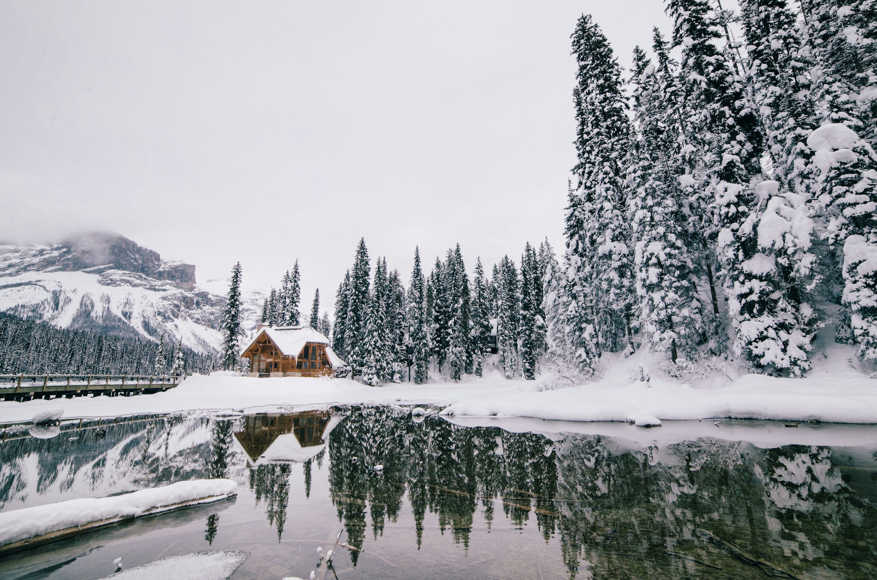 Snow-covered cabin beside a frozen lake, with pine forest and mountains reflected in the water