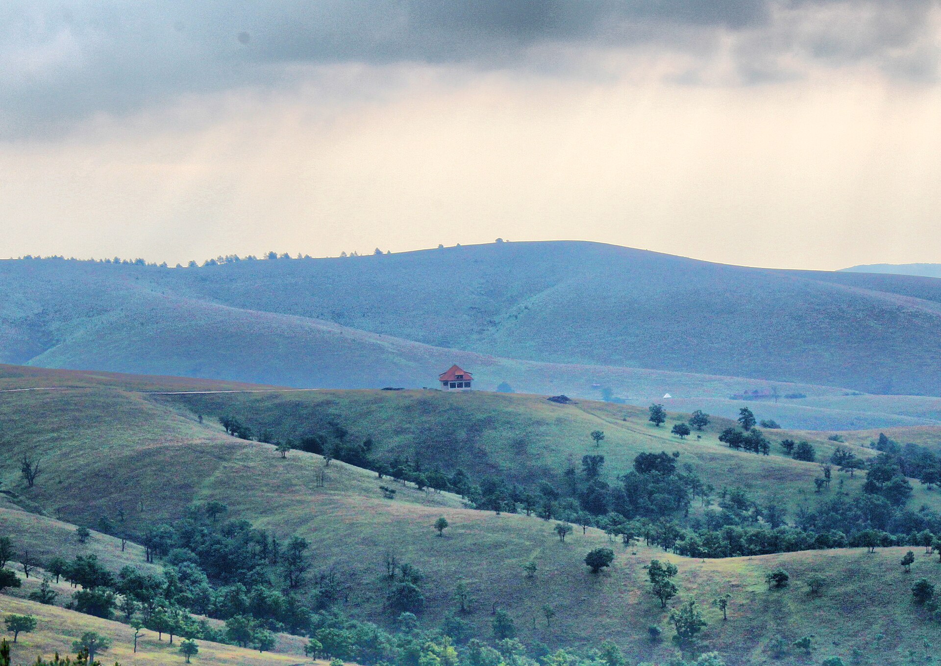 Rolling grassy hills with scattered trees and a small house under storm clouds in Zlatibor